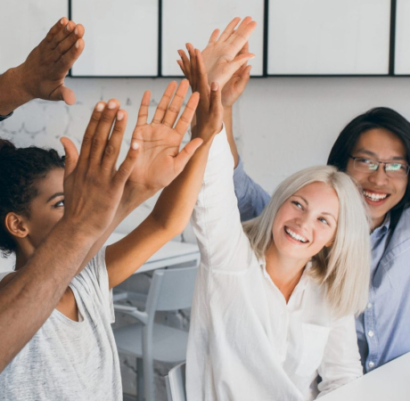 Adorable girl with short blonde hairstyle slapping hands with african female colleague. Indoor photo of joyful coworkers celebrating beginning of vacation at workplace.